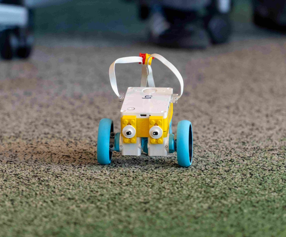 Ein Lego-Roboter mit gelben und blauen Rädern, der auf einem Teppichboden fährt. Er ist für Programmierprojekte im IT-Bereich konzipiert.