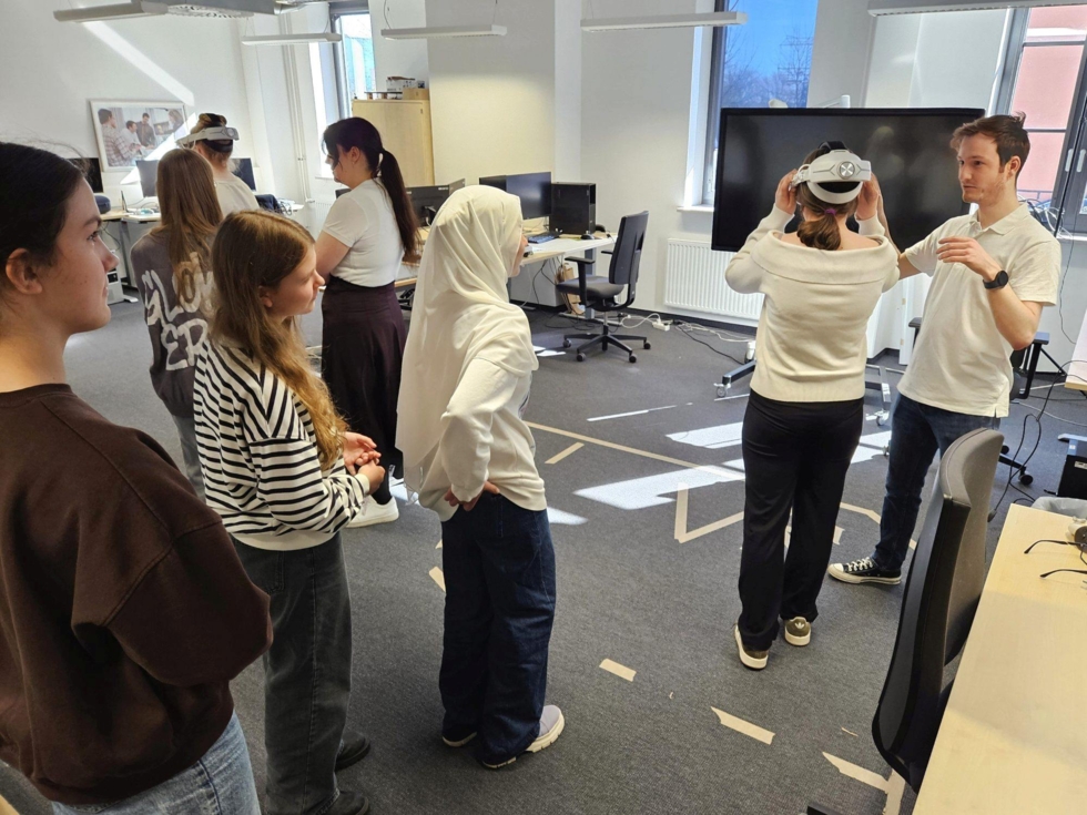 Gruppe von Girls' Day-Teilnehmerinnen in einem Büro, eine Teilnehmerin trägt eine VR-Brille, ein Mitarbeiter erklärt etwas, andere beobachten aufmerksam