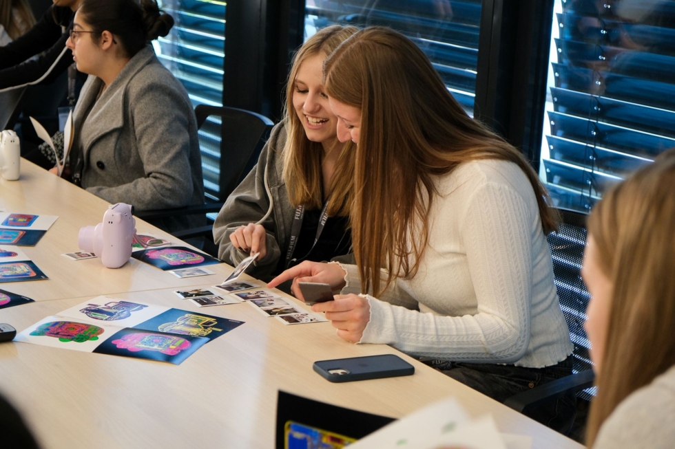 Zwei Girls' Day-Teilnehmerinnen sitzen an einem Tisch und betrachten Fotos. Im Hintergrund sind weitere Girls' Day-Teilnehmerinnen zu sehen