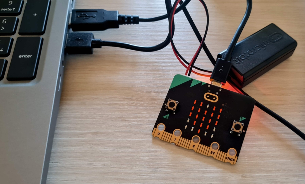Ein Mini-Computer mit mehreren angeschlossenen Kabeln, der an einen Laptop angeschlossen ist. Auf dem Mini-Computer sind Tasten und eine LED-Anzeige sichtbar.