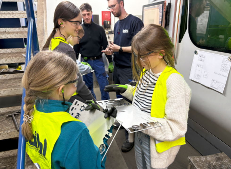 Die Girls' Day-Teilnehmerinnen halten ein Metallteil, während die Mitarbeiter Anweisungen geben