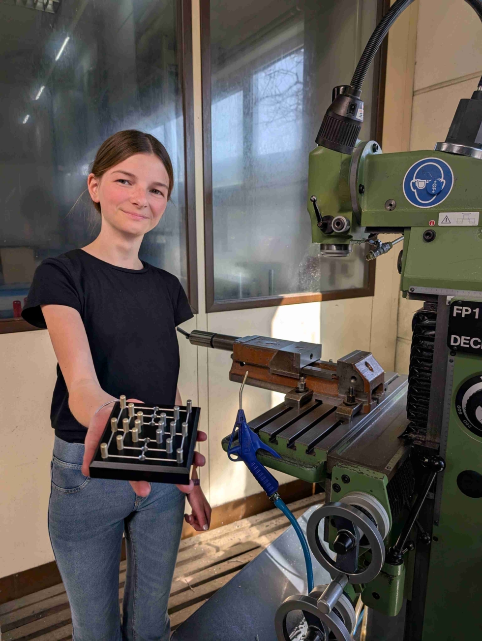 Girls'Day-Teilnehmerin steht lächelnd vor einer Fräsmaschine und hält ein selbstgefertigtes Werkstück in der Hand