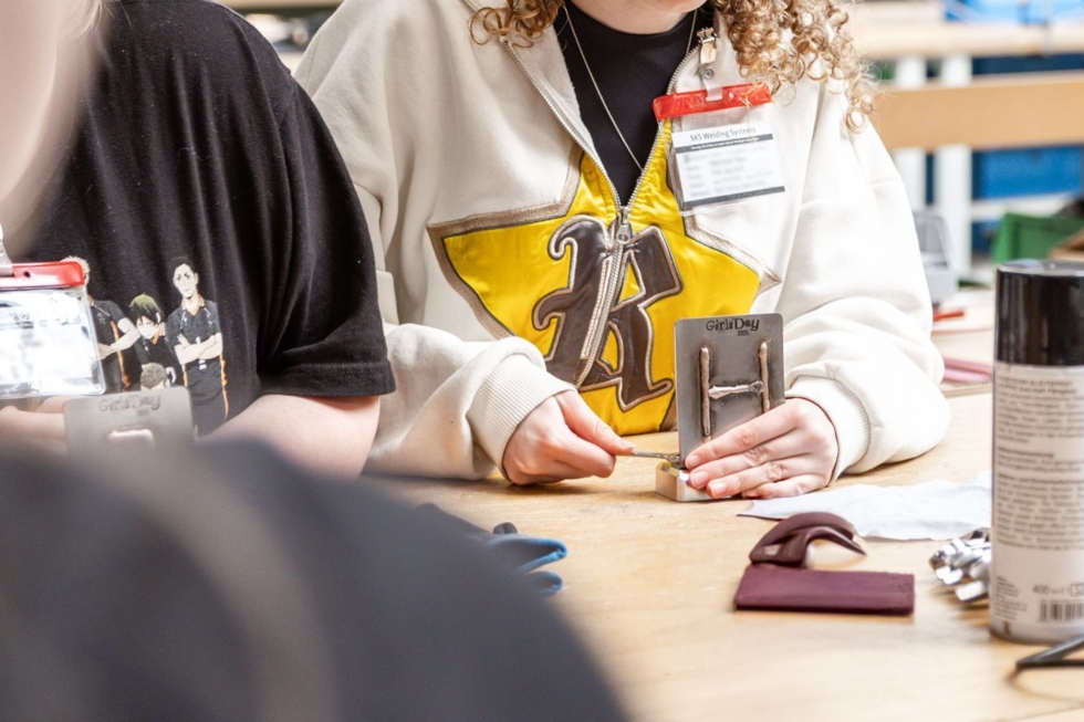 Girls' Day-Teilnehmerin arbeitet an einem Maschinenbauprojekt, trägt einen weißen Hoodie mit gelbem Logo, umgeben von Werkzeugen und Materialien