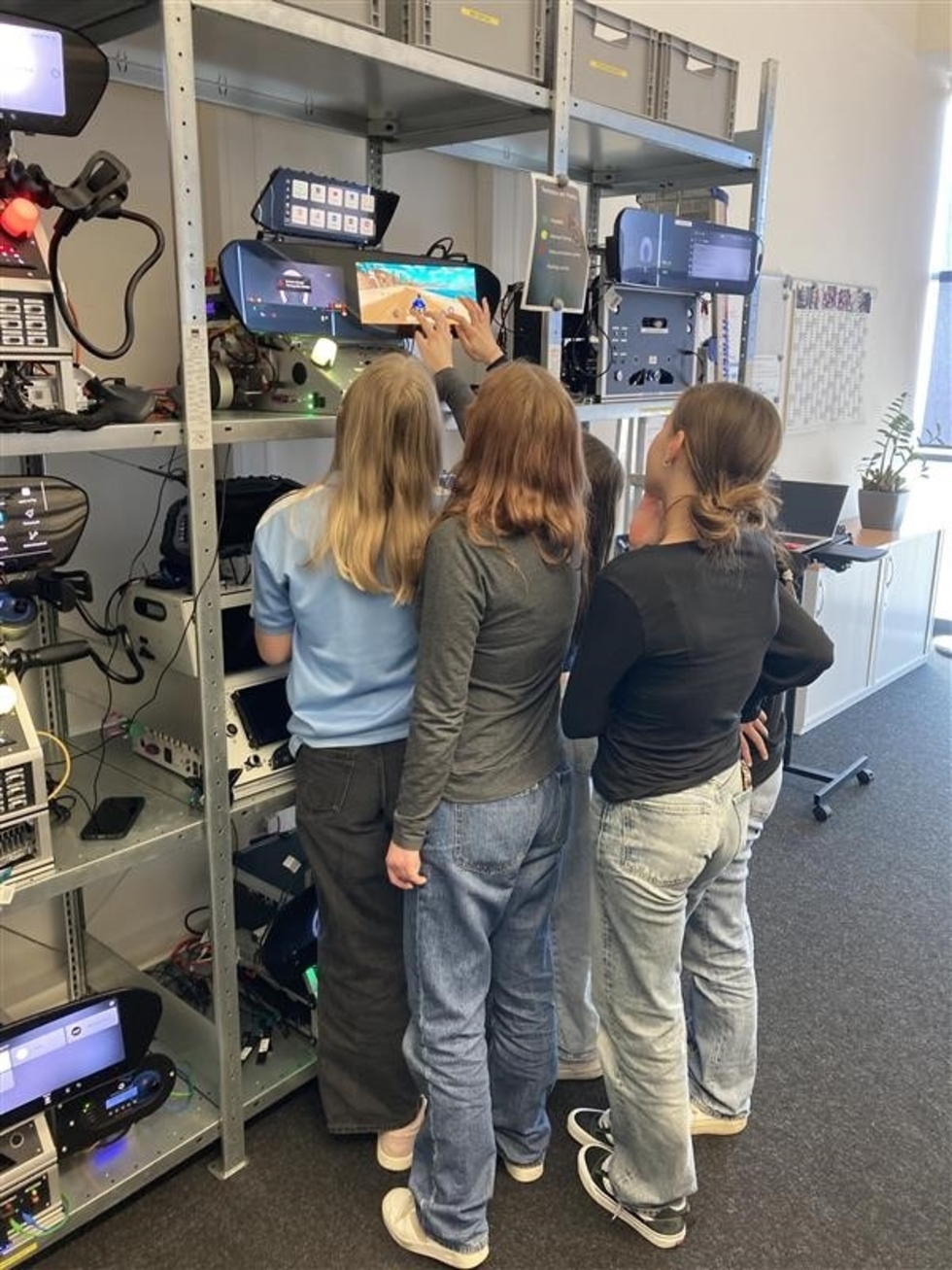 Drei Girls' Day-Teilnehmerinnen stehen vor einem Regal mit Beleuchtungstechnik und betrachten ein beleuchtetes Gerät an der oberen Regalebene