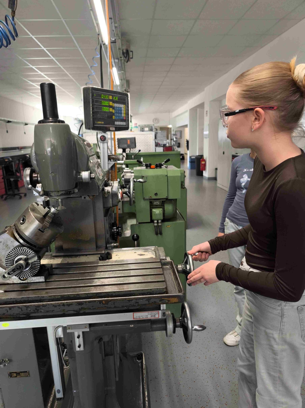 Eine junge Frau bedient eine Maschine in einer Werkstatt. Sie trägt eine Schutzbrille und hat einen konzentrierten Gesichtsausdruck.