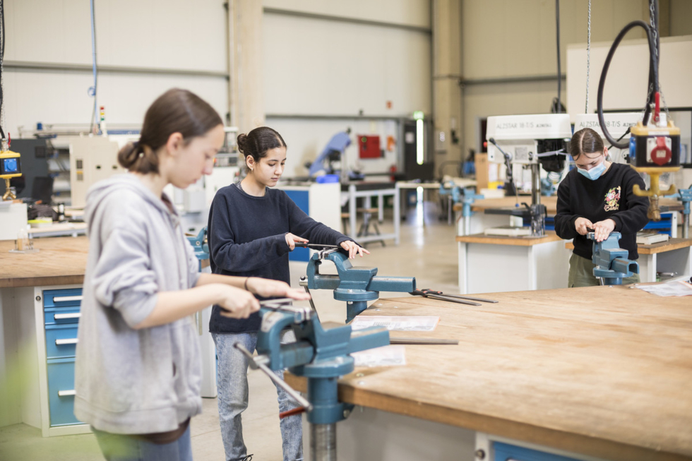 Drei Mädchen feilen an Metallplatten, die in einen Schraubstock eingespannt sind.