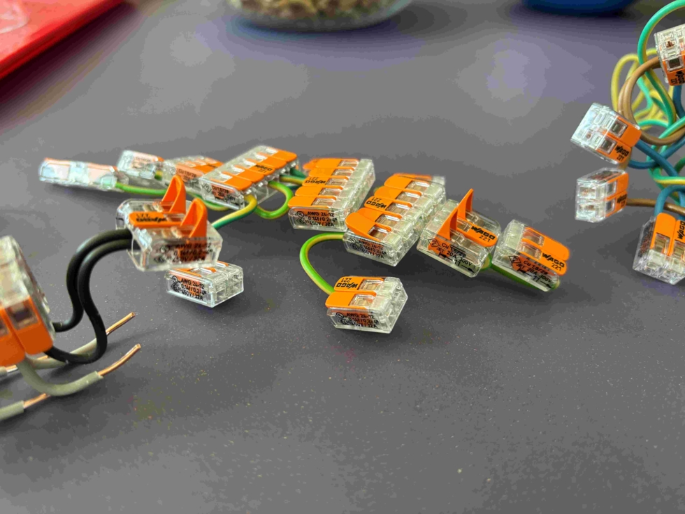 Mehrere transparente Verbindungsklemmen mit orangefarbenen Markierungen auf einem Tisch, umgeben von elektrischen Drähten