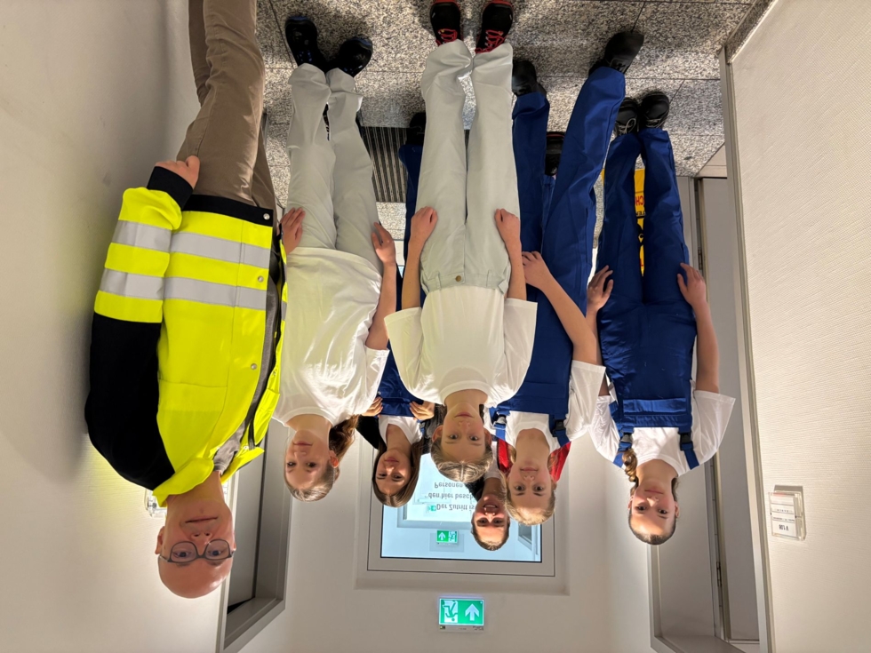 Sechs Girls' Day Teilnehmerinnen in Arbeitskleidung stehen in einem Flur. Der Mitarbeiter trägt eine gelbe Warnjacke