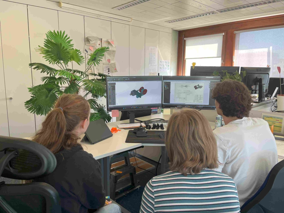 Zwei Girls' Day-Teilnehmerinnen und ein Mitarbeiter sitzen an einem Schreibtisch und betrachten zwei Monitore mit technischen Zeichnungen in einem Büro