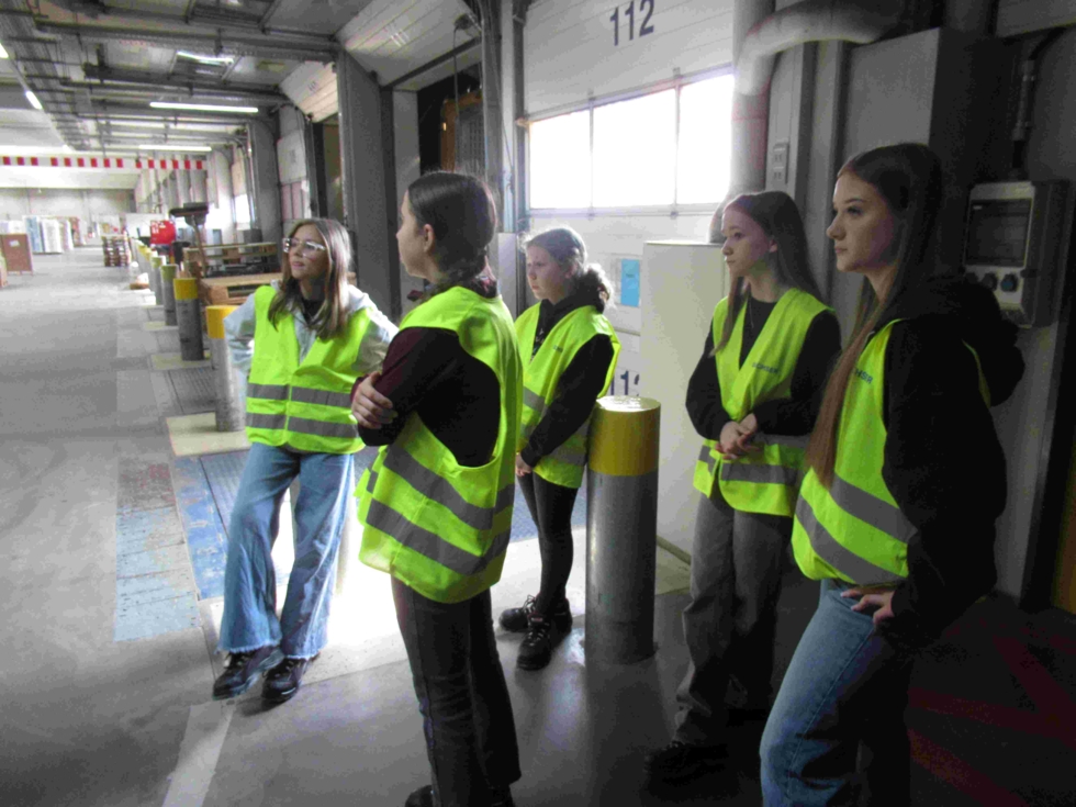 Fünf Girls' Day-Teilnehmerinnen stehen in gelben Sicherheitswesten in einer Lagerhalle und hören aufmerksam zu