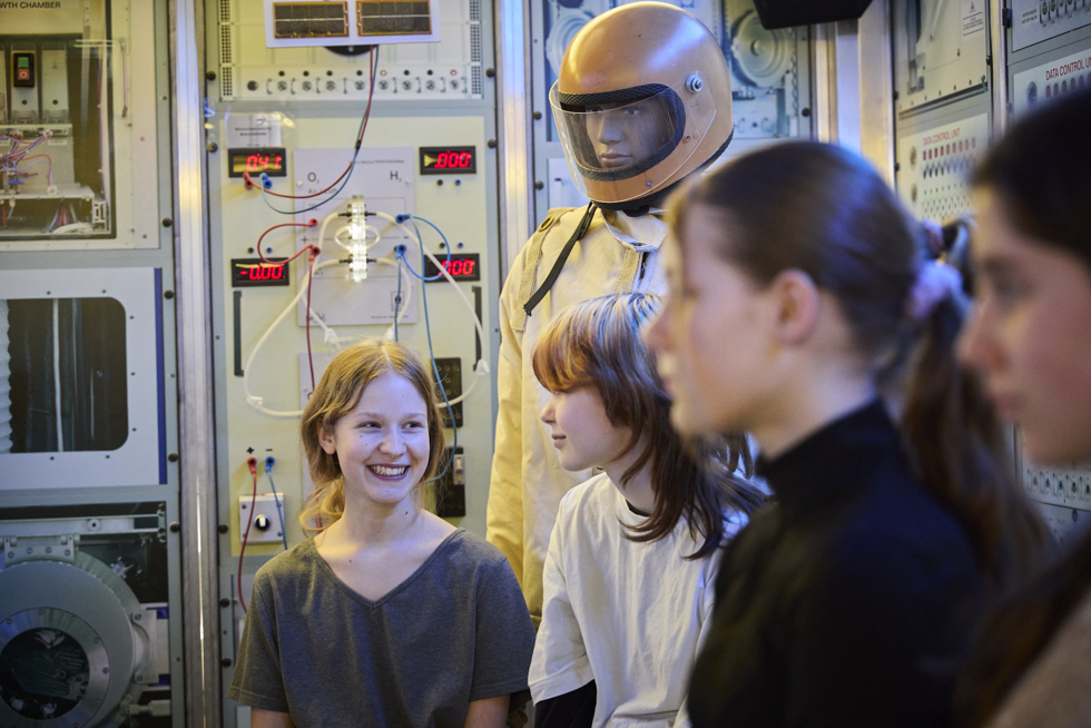 Girls'Day-Teilnehmerinnen sitzen vor Schaltaschränken, im Hintergrund steht eine lebensgroße Puppe mit Raumanzug.