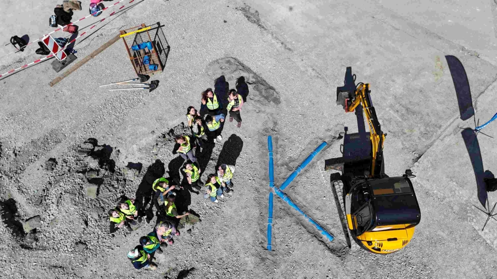 Luftaufnahme einer Baustelle mit den Girls' Day-Teilnehmerinnen in gelben Sicherheitswesten, die ein großes 'K' aus blauen Rohren bilden. Ein Bagger steht daneben.