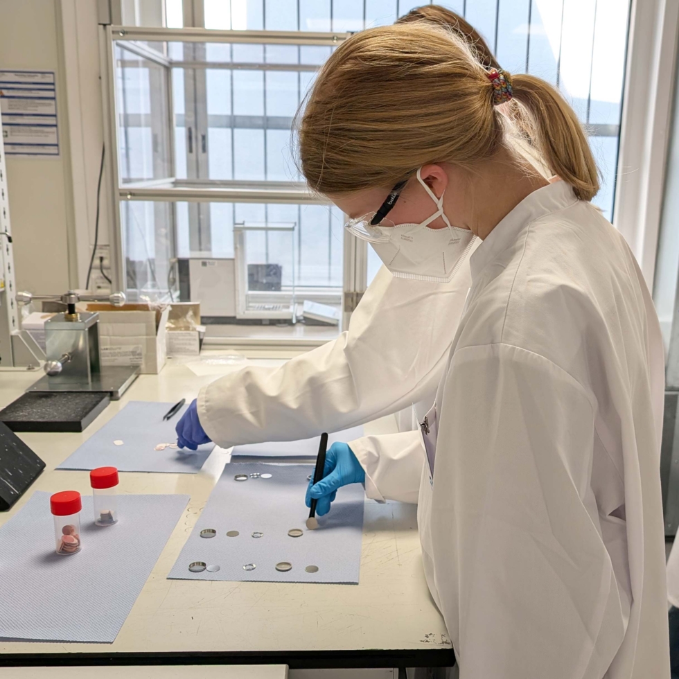 Girls' Day-Teilnehmerin in Laborkleidung untersucht Proben auf einem Tisch im Labor, umgeben von wissenschaftlichen Materialien.
