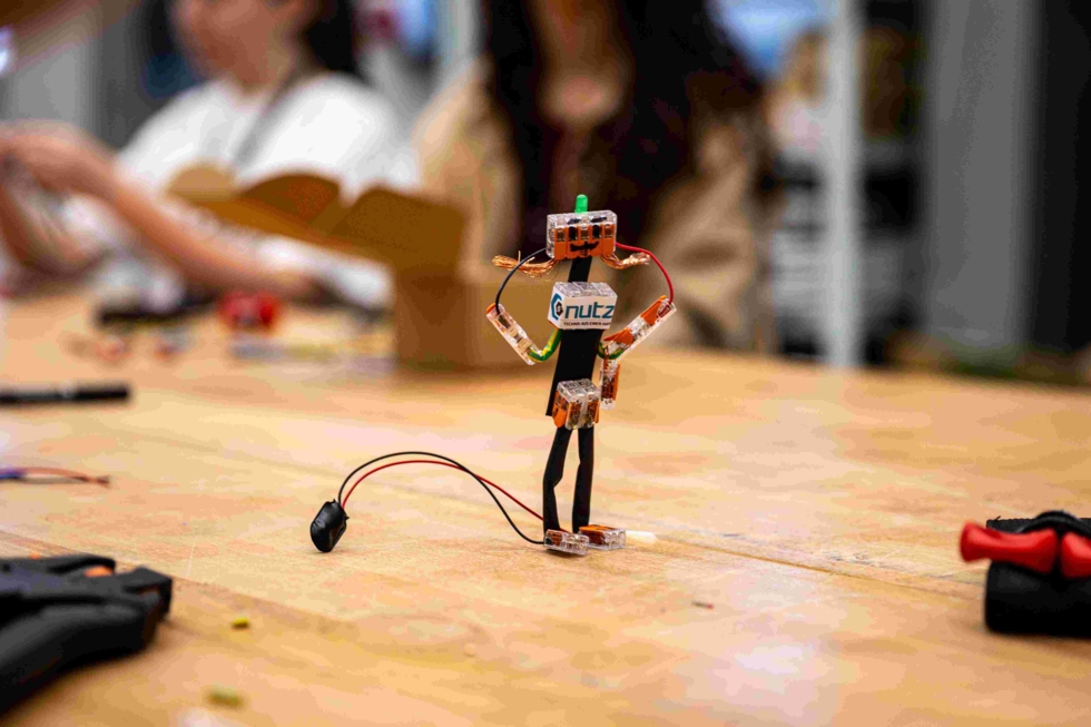 Ein selbstgebauter Roboter aus elektrischen Bauteilen steht auf einem Tisch in einer Elektrowerkstatt. Im Hintergrund sind weitere Girls' Day-Teilnehmerinnen beim Basteln zu sehen