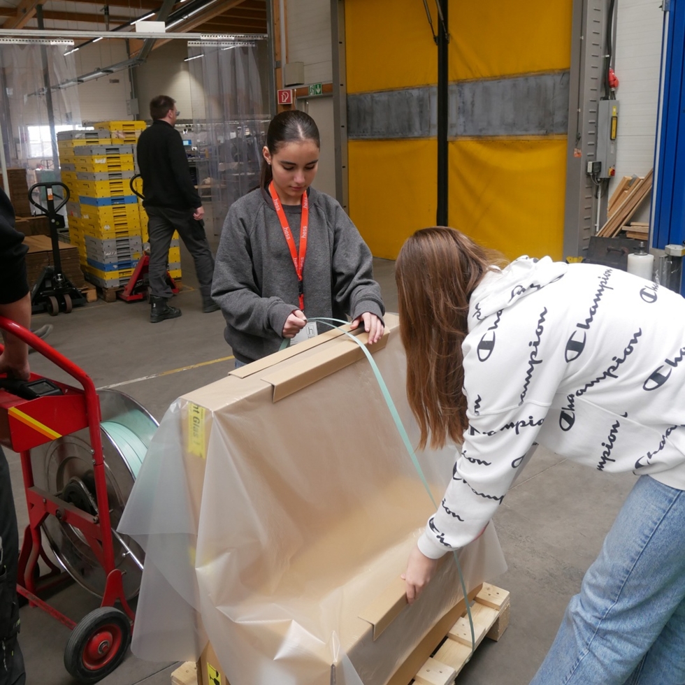 Zwei Girls' Day-Teilnehmerinnen verpacken in einer Lagerhalle ein großes Paket mit braunem Papier und Klebeband