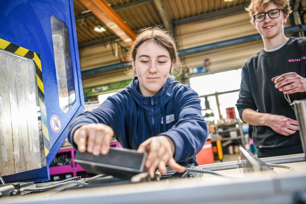 Eine junge Frau in blauer Arbeitskleidung bedient eine Maschine in einer Werkstatt, während ein lächelnder junger Mann im Hintergrund zuschaut.