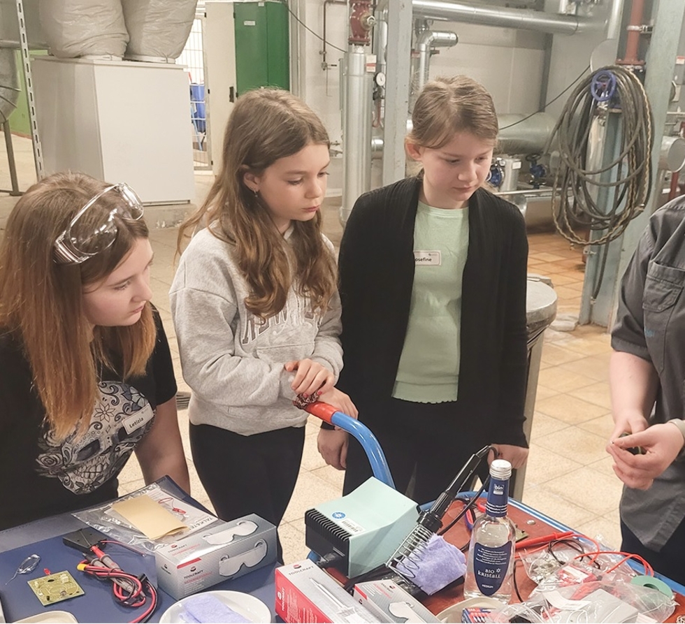 Die drei Girls' Day-Teilnehmerinnen beobachten einen Mitarbeiter, der ihnen technische Geräte erklärt. Im Hintergrund sind Rohre und Maschinen sichtbar.