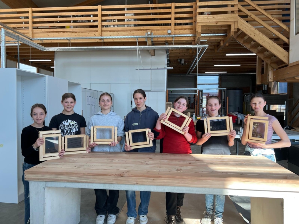 Sechs Teilnehmerinnen des Girls' Day halten stolz ihre Holzrahmen mit bearbeiteten Ergebnissen in einer Werkstatt