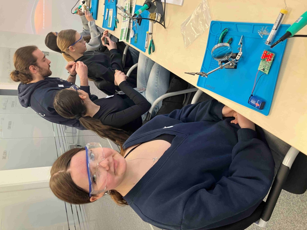 Girls' Day Teilnehmerinnen sitzen am Tisch und erproben sich beim Löten einer elektronischen Schaltung unter Anleitung des Mitarbeiters