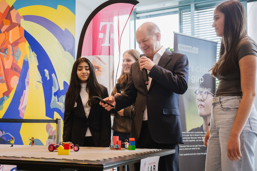 Drei Schülerinnen beobachten Olaf Scholz dabei, wie er ein Fahrzeug fernsteuert