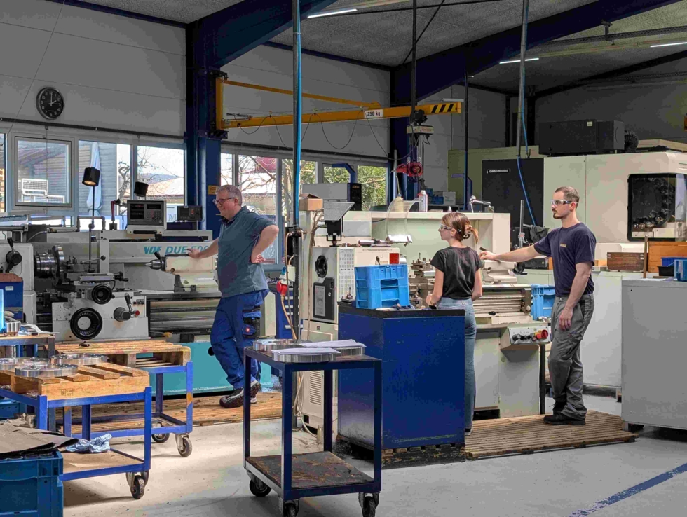 Girls' Day-Teilnehmerin und zwei Mitarbeiter in einer Werkstatt. Sie arbeiten an Maschinen wie Dreh- und Fräsmaschinen.