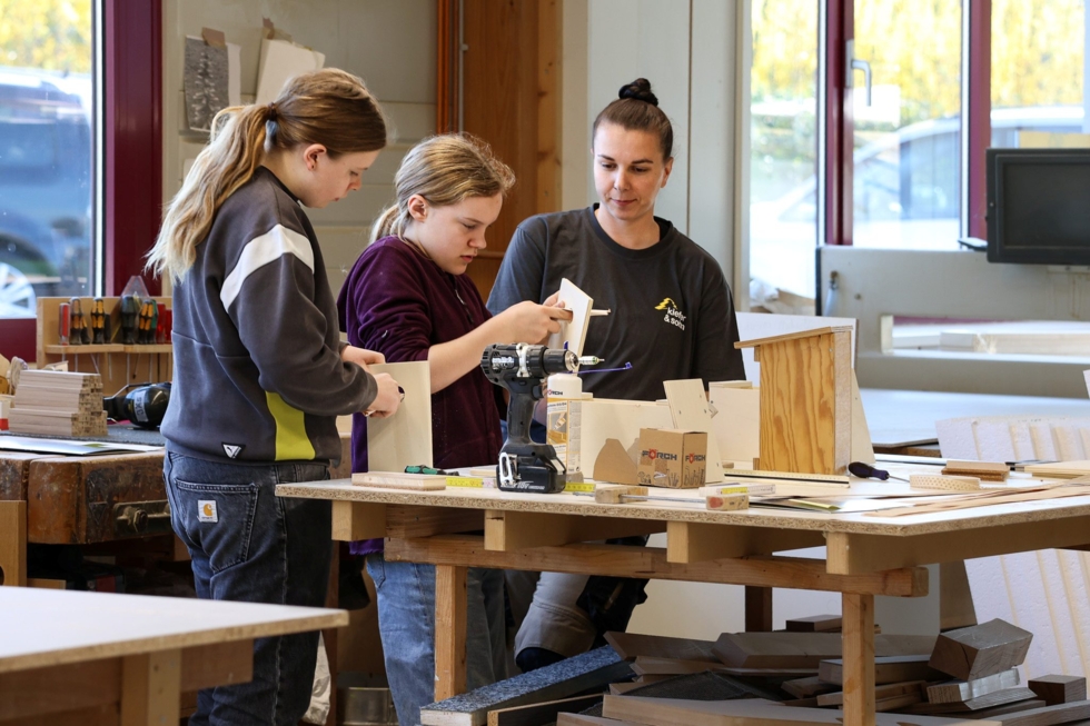 Girls' Day-Teilnehmerinnen und eine Auszubildende arbeiten an einem Tisch in einer Werkstatt, umgeben von Holzmaterialien und Werkzeugen