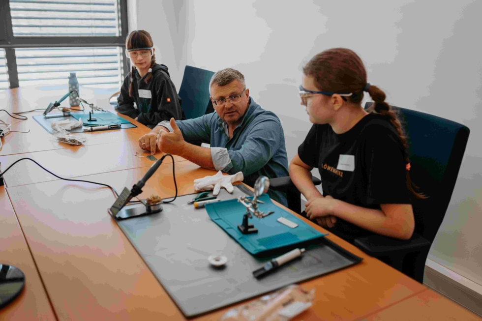 Girls' Day Teilnehmerinnen sitzen am Tisch mit einem Mitarbeiter und machen eine Lötübung