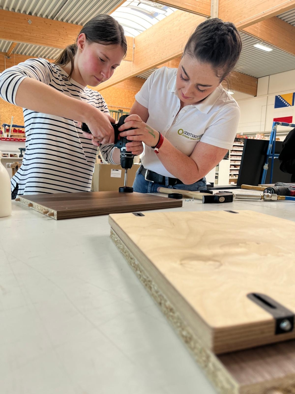 Zwei Frauen arbeiten gemeinsam an einem Holzprojekt, eine zeigt der anderen, wie man ein Werkzeug benutzt.