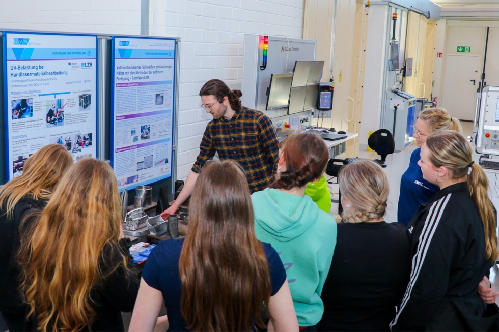 Mitarbeiter zeigt Girls' Day-Teilnehmerinnen verschiedene technische Bauteile, während im Hintergrund Infotafeln und Maschinen zu sehen sind