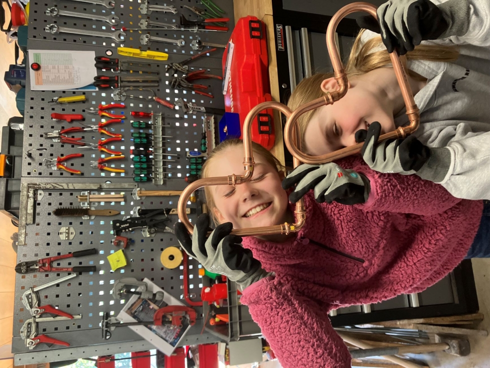 Zwei Girls' Day-Teilnehmerinnen präsentieren selbst gebastelte Rohrherzen in einer Werkstatt, umgeben von Werkzeugen