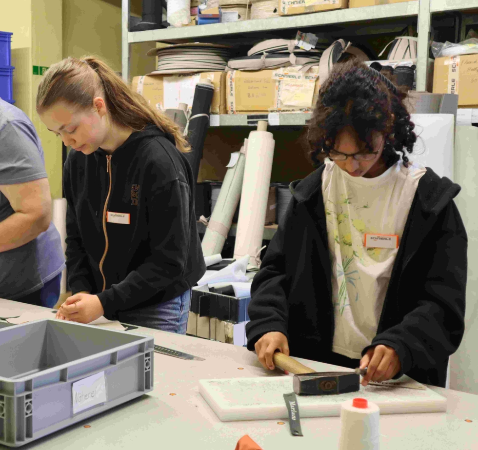 Zwei Girls' Day-Teilnehmerinnen arbeiten konzentriert an einem Tisch in einem Lagerraum, umgeben von Materialien und Werkzeugen