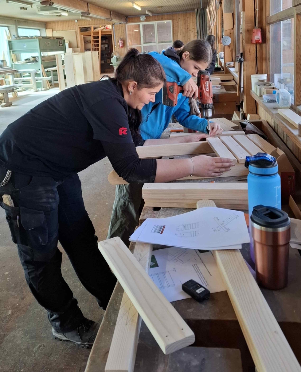 Zwei Frauen arbeiten in einer Holzwerkstatt, eine hält Holzstücke fest, die andere bearbeitet sie. Werkzeuge und Pläne sind auf dem Tisch.