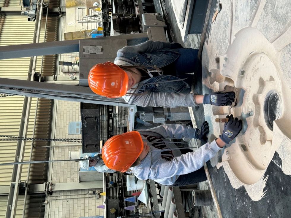 Zwei Girls' Day-Teilnehmerinnen arbeiten in Schutzhelmen und Handschuhen an einer großen Maschinenkomponente in einer Fabrik