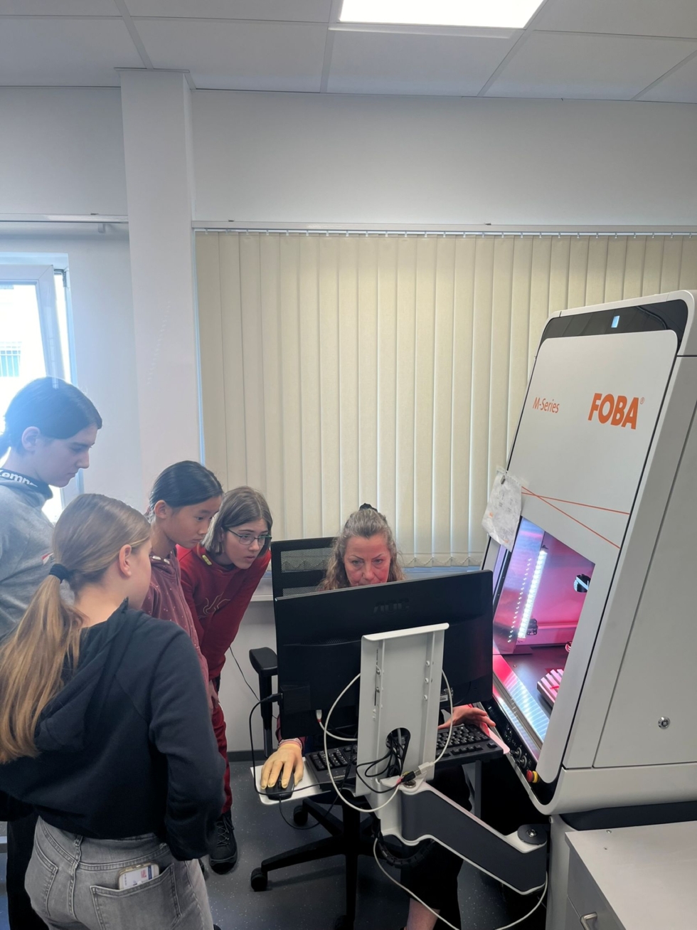 Fünf Girls' Day-Teilnehmerinnen stehen um einen Computer in einem Labor. Eine Mitarbeiterin sitzt am Computer und arbeitet mit einer Maschine