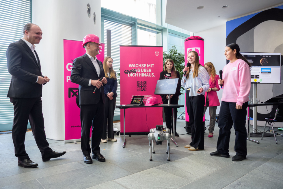 Schülerinnen präsentieren dem Bundeskanzler einen Roboterhund und erläutern den Einsatz von Künstlicher Intelligenz. Im Hintergrund sind pinke Banner mit Telekom-Logos und Schriftzügen zu sehen.