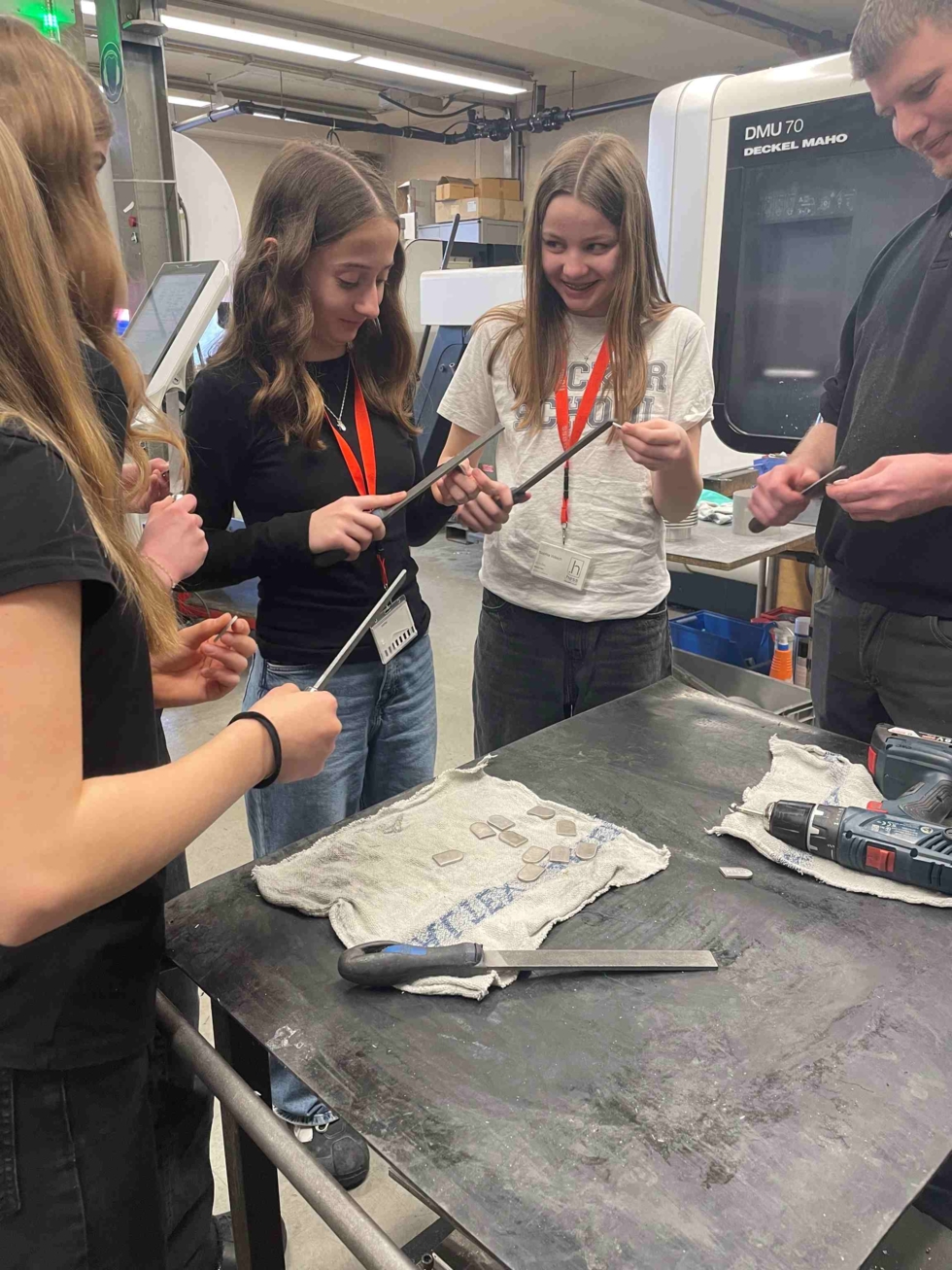 Mitarbeiter zeigt Girls' Day-Teilnehmerinnen in Werkstatt den Umgang mit Werkzeugen an einem Arbeitstisch mit Tüchern und Maschinen im Hintergrund