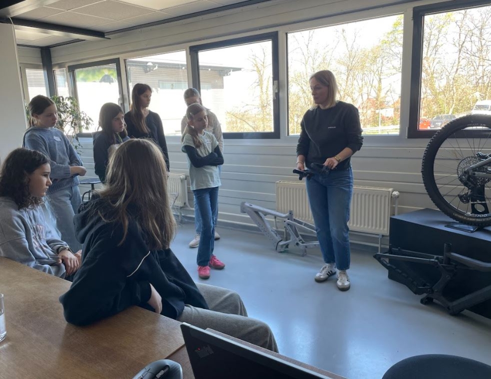 Mitarbeiterin erklärt einer Girls' Day-Teilnehmerinnen in einem hellen Raum mit großen Fenstern technische Details an einem Fahrrad auf einer Hebebühne